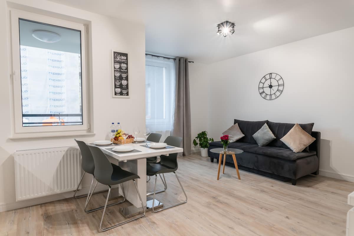 Una sala moderna con comedor, un sofá gris, un reloj en la pared y una ventana con cortinas.