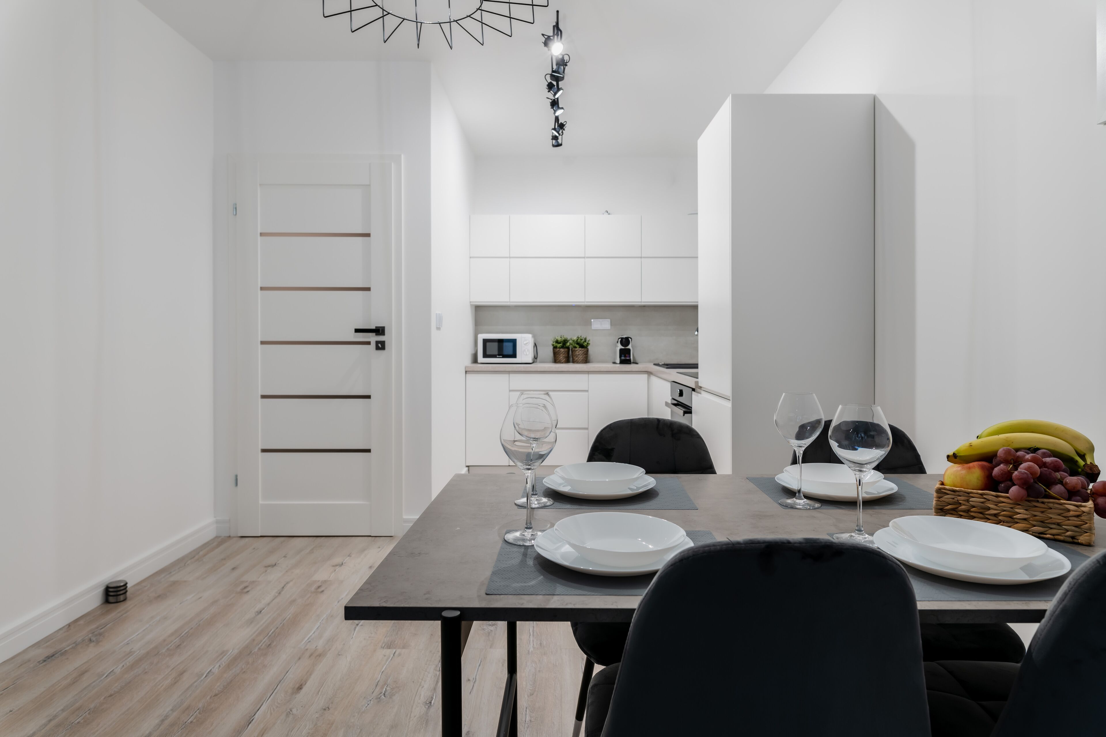 A modern dining area with a wooden floor, white walls, and a kitchen in the background. There are two dining tables set with plates and wine glasses.