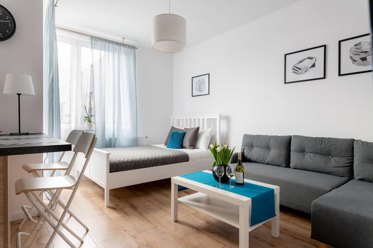 Una sala de estar moderna y bien iluminada con una cama, un sofá gris, un comedor con mesa y sillas, y obras de arte en las paredes.