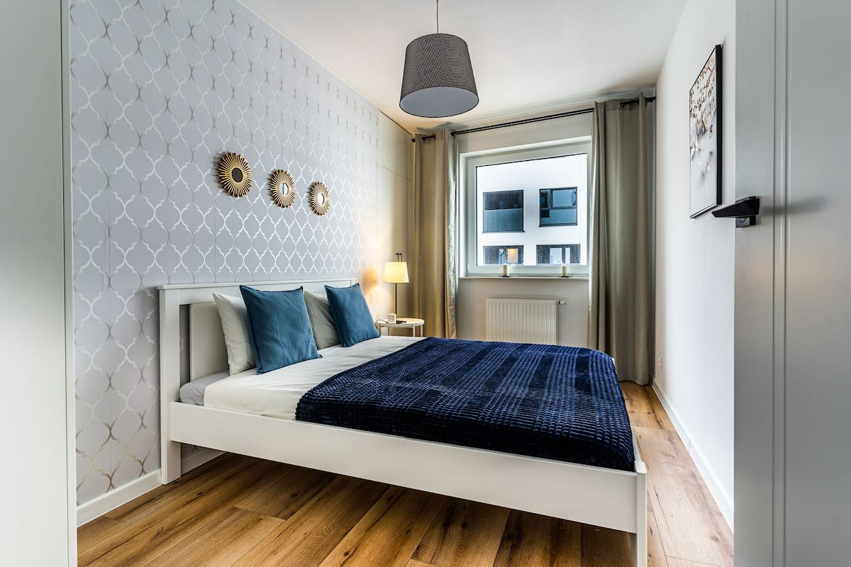 Un dormitorio moderno con una cama blanca, papel tapiz estampado y vistas a edificios a través de una ventana.