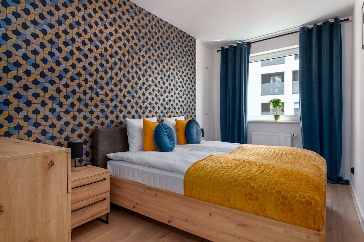 Un dormitorio moderno con pared de estampado geométrico, estructura de cama de madera y colcha amarilla con textura.