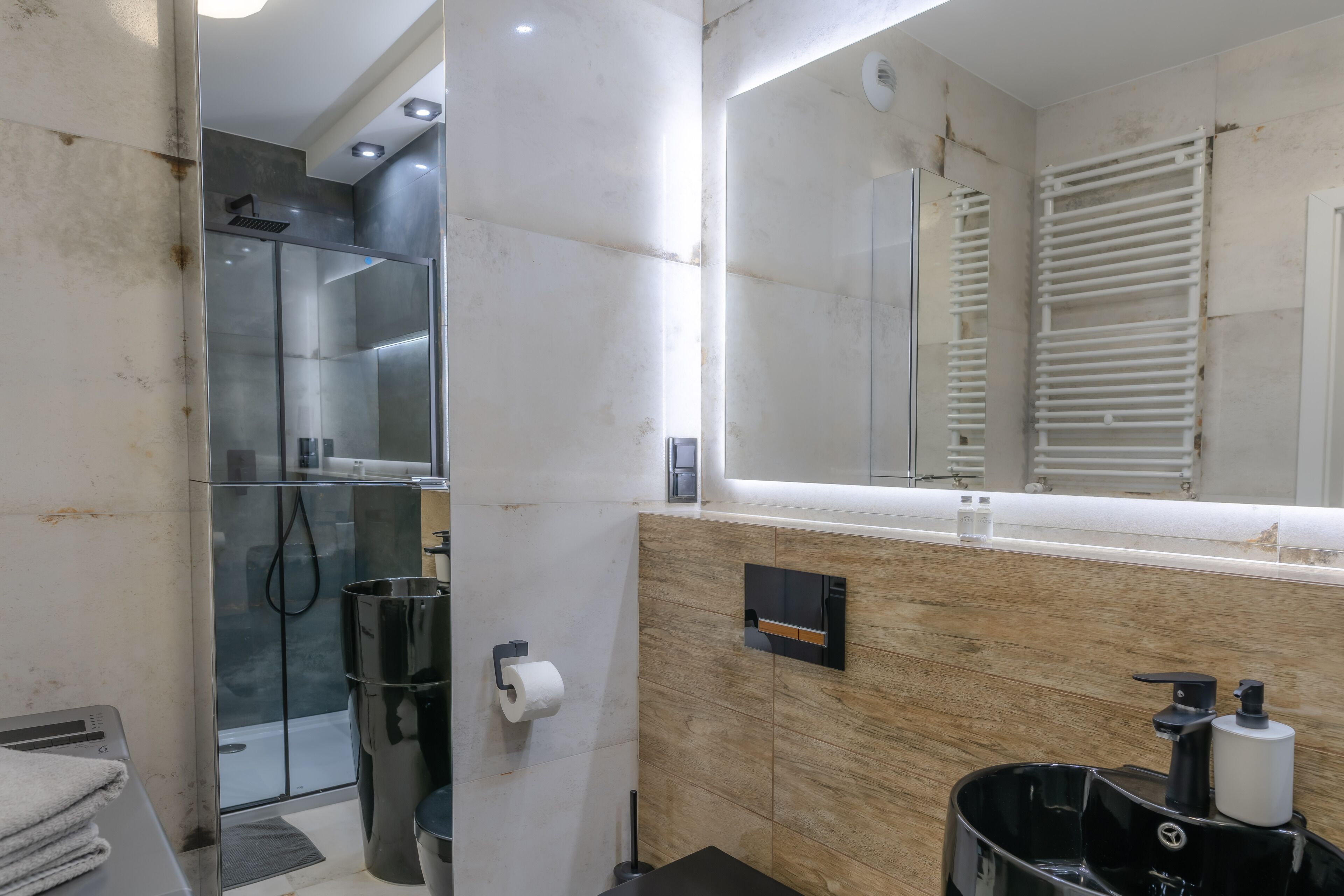 Un baño moderno con ducha de vidrio, dispensador de papel higiénico y un cubo de basura negro.