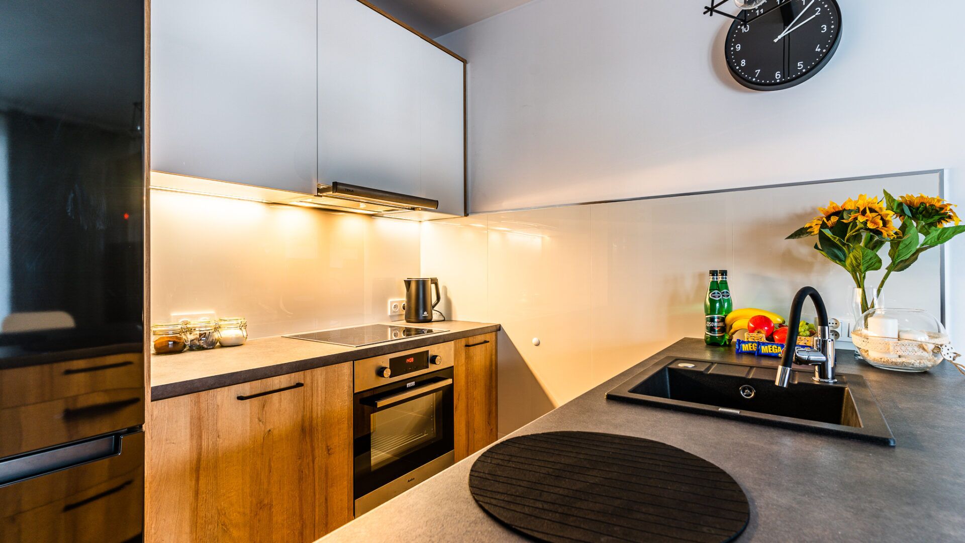 Een moderne keuken met houten kastjes, een inbouwoven, een koffiezetapparaat en een klok aan de muur.