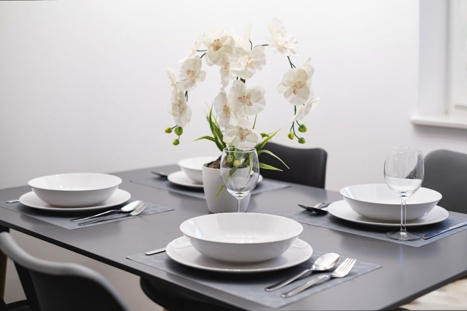 A dining table set with white plates, bowls, and cutlery, accompanied by a vase of white flowers.