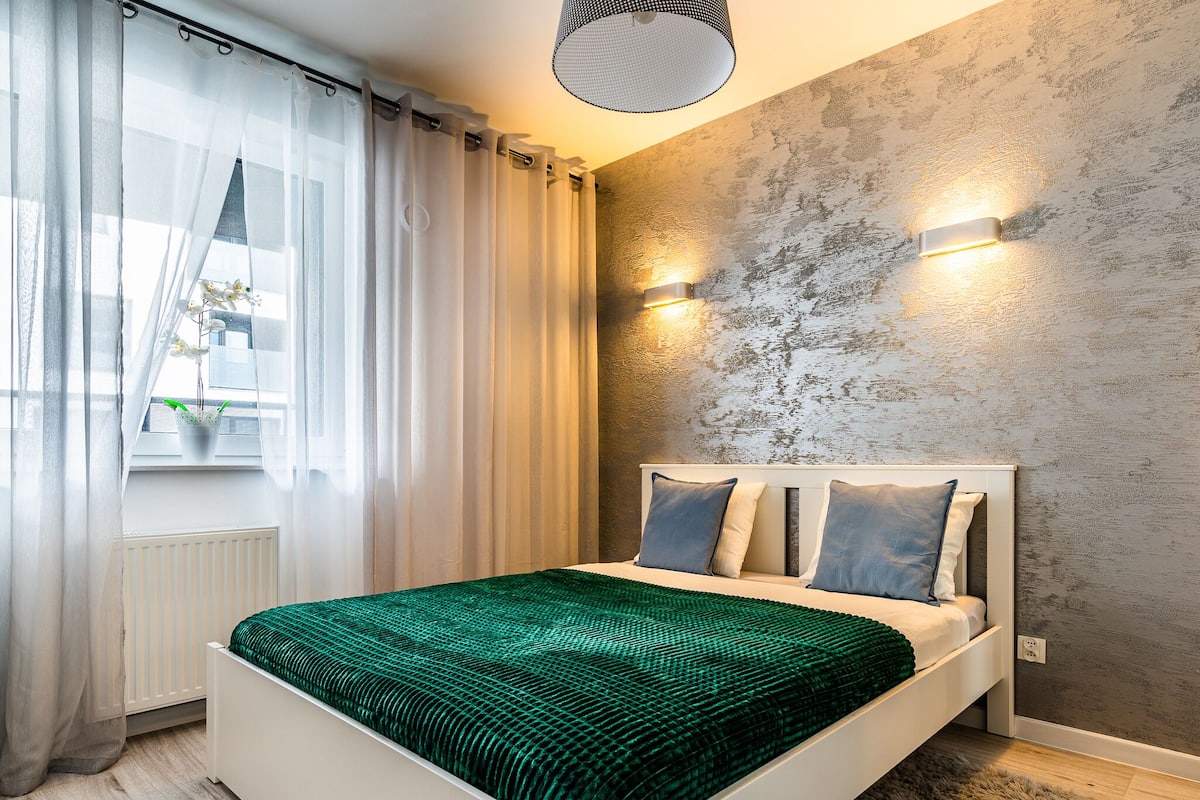Un dormitorio moderno con cama, cortinas, una ventana con planta y una pared texturizada.