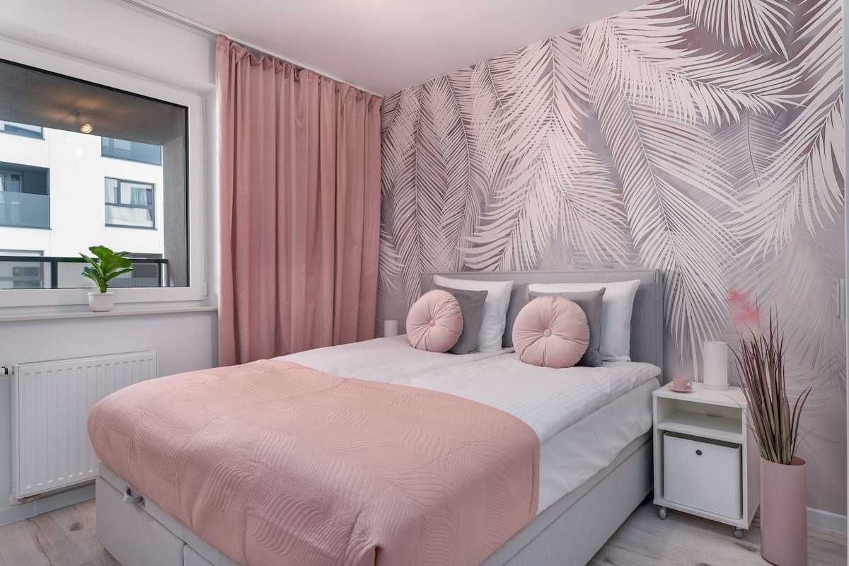 Un dormitorio con cama, mesita de noche, ventana con cortinas, radiador y un papel tapiz con diseño de hojas.