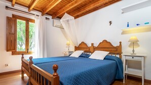 Un dormitorio con estructura de cama de madera, colcha acolchada azul, dos mesitas de noche con lámparas y una ventana con cortinas blancas.