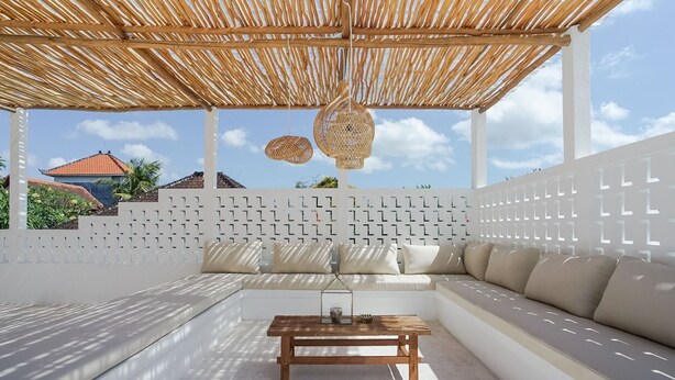 A covered outdoor seating area with a wooden table, white cushions, and a woven hanging light fixture. - Avanoa Villa (Canggu)