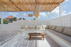 Un área de estar al aire libre techada con una mesa de madera, cojines blancos y una lámpara colgante tejida.