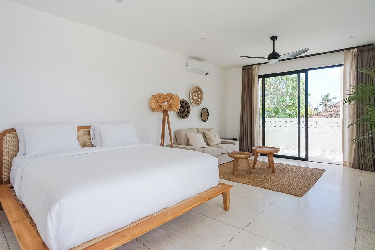 A modern bedroom with a large bed, a ceiling fan, a sofa, and a view of the outdoors.