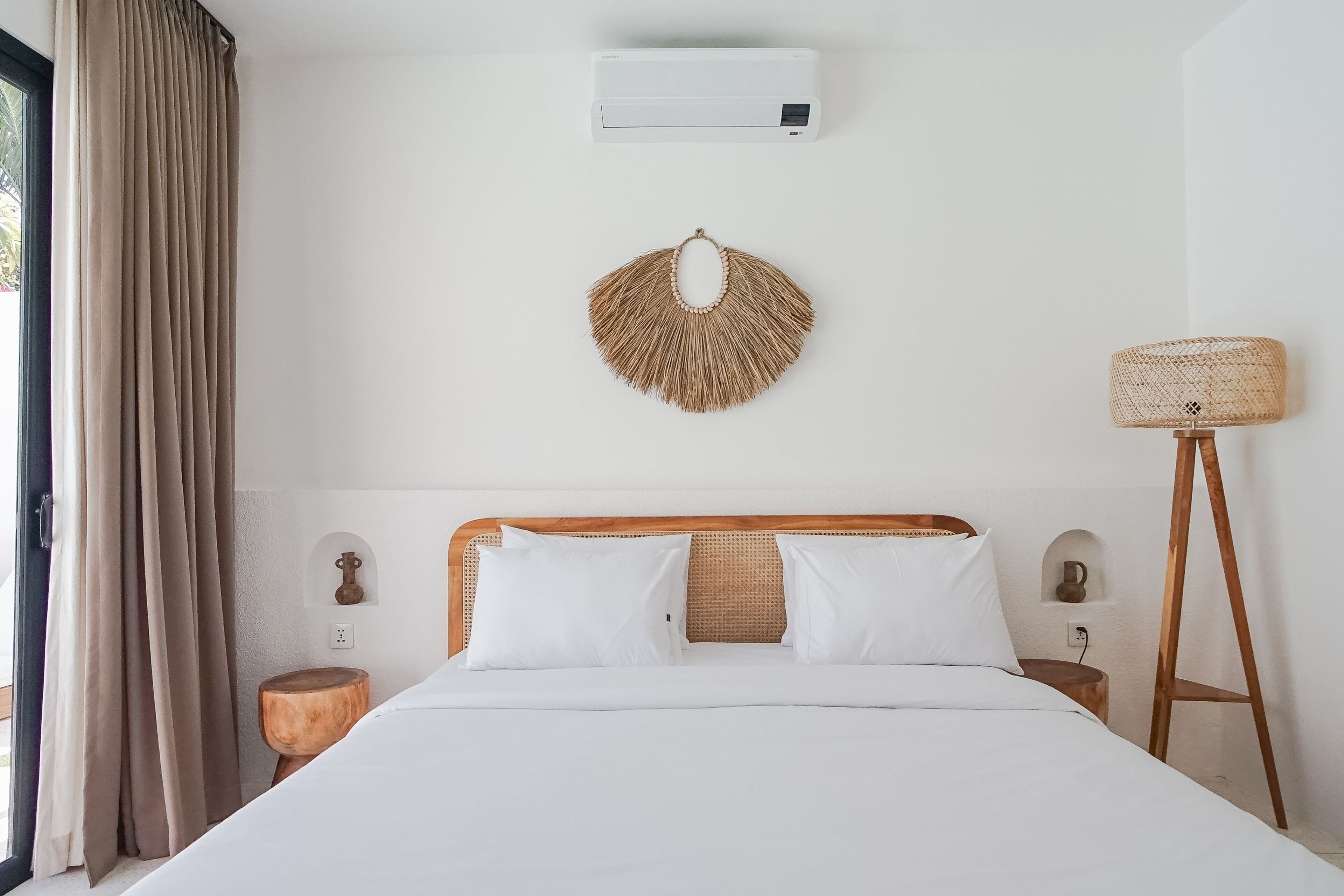Ein ordentlich eingerichtetes Schlafzimmer mit Holz-Kopfstütze, weißer Bettwäsche, Wand-Klimaanlage, Wandteppich aus Gewebe, einer Nachttischlampe und einem kleinen Holzschemel.