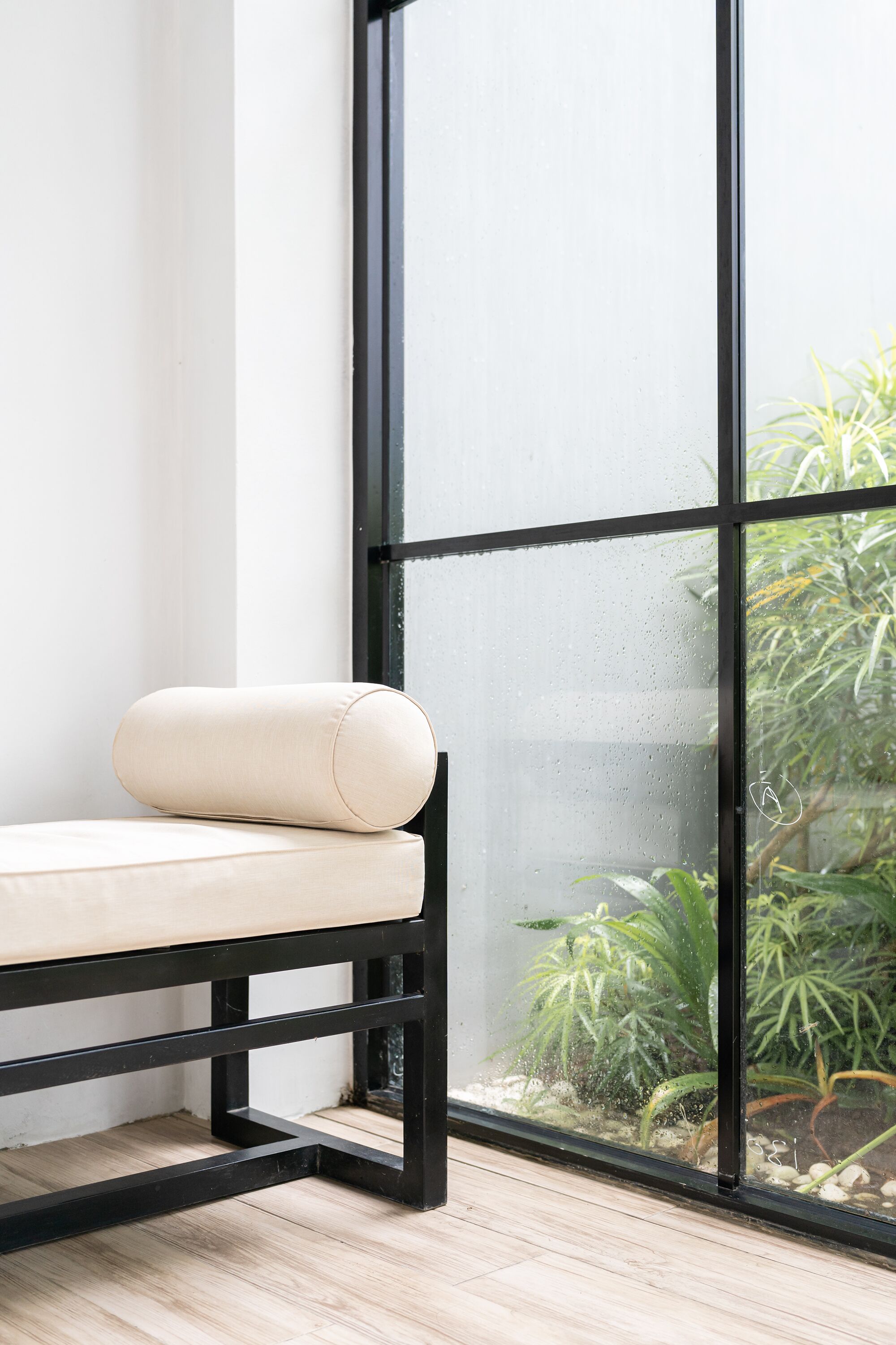 A corner of a room with a black metal bench and a cushion, a large window with a view of indoor plants, and a wooden floor.