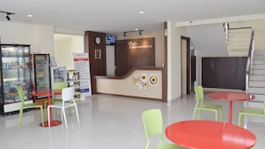 A hotel reception area with a reception desk, a clock, a TV screen, and a refrigerator. There are green and red tables and chairs. - Homestay 82 Syariah Palembang (Palembang)