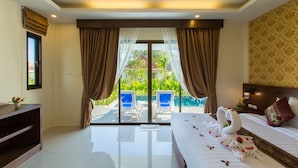 Habitación de hotel con cama, mesita de noche, televisor, ventana con cortinas y vistas a una piscina y vegetación, además de un cabecero con un cojín decorativo.