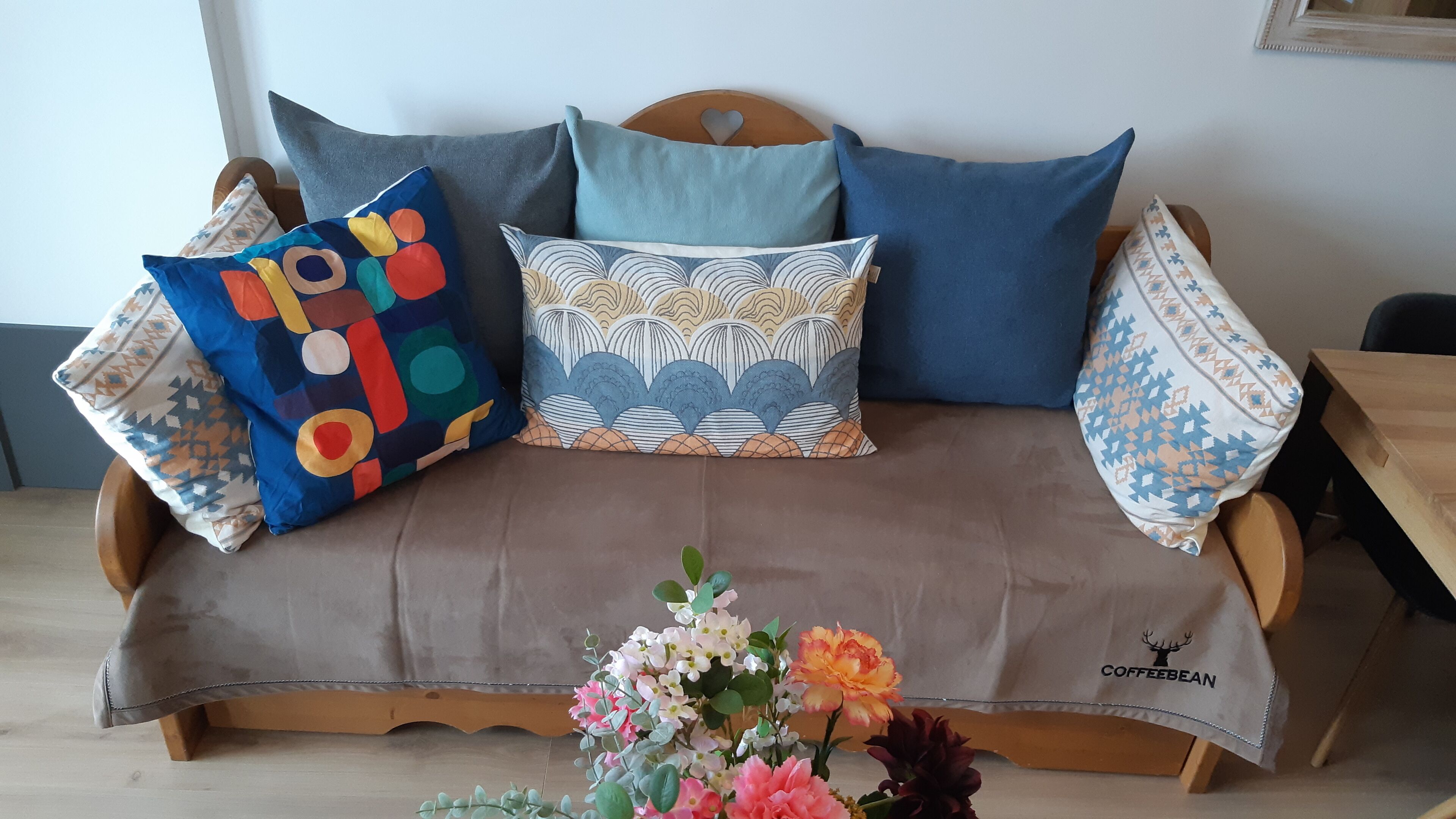 A wooden sofa with a brown cover, adorned with colorful and patterned pillows, and a floral arrangement on the floor.