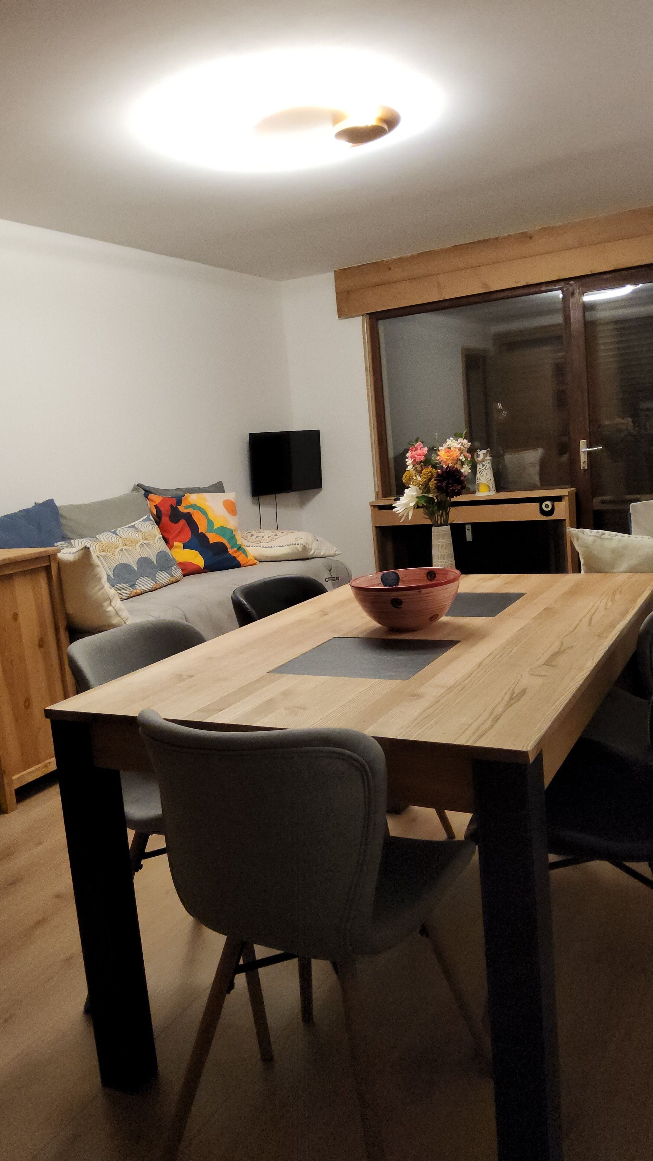 A dining area with a wooden table, two grey chairs, a TV on the wall, and a sofa with colorful cushions.