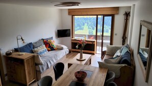 Un salon confortable avec un canapé, une table basse et une table à manger. Un téléviseur est fixé au mur et une fenêtre offre une vue sur l’extérieur.