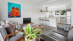 A modern living room with a sofa, a coffee table, and a TV. There is a kitchen area with a dining table and chairs.