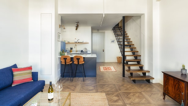 Een moderne woonkamer met een blauwe bank, een salontafel met een fles wijn, en een keukenhoek met een barkruk. Een houten trap leidt naar een verdieping.