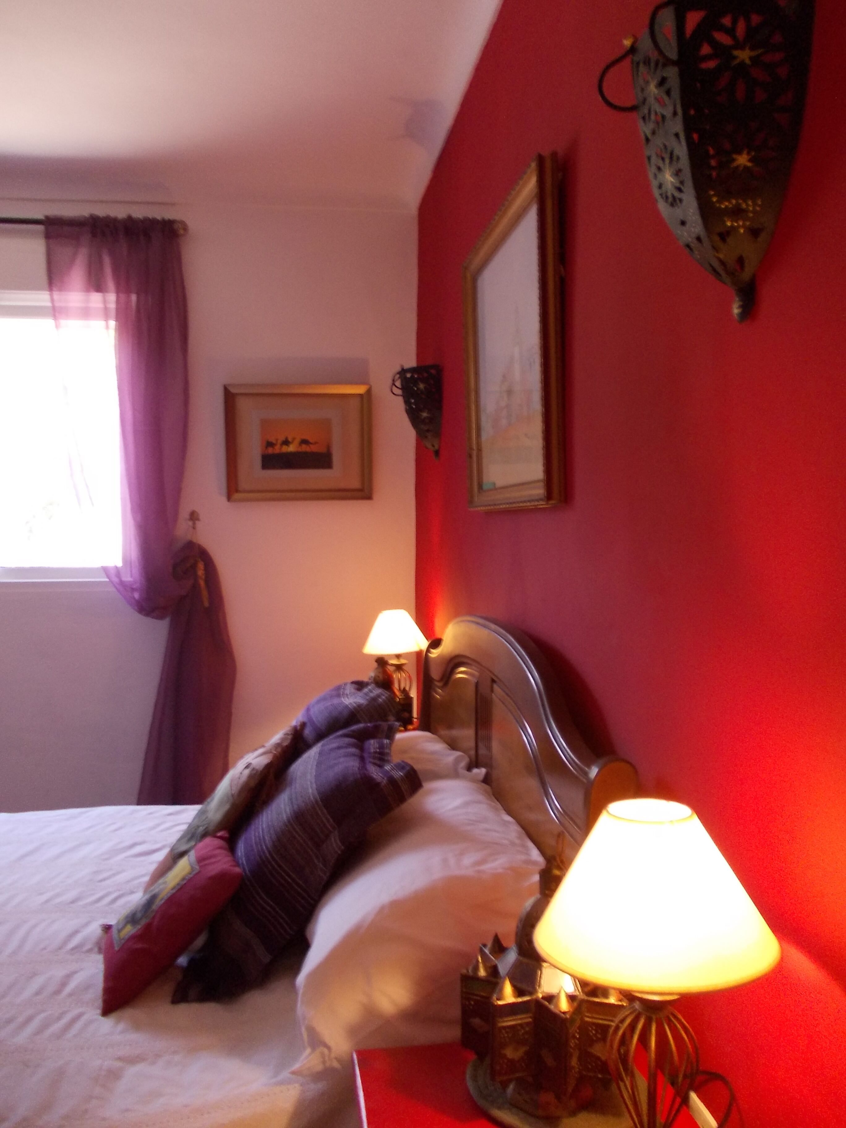 A bedroom with a bed, two bedside lamps, a window with purple curtains, and a framed picture on the wall.