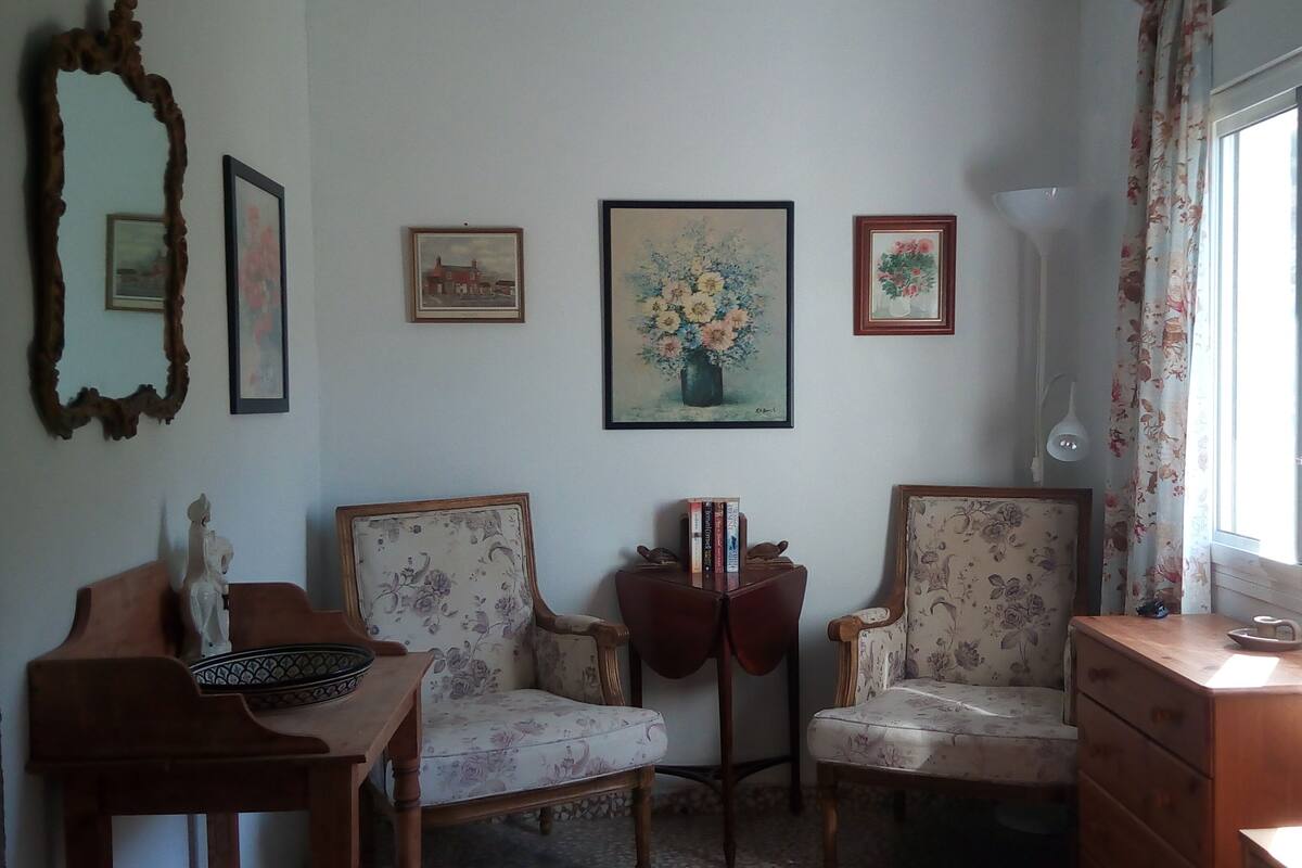 A room with two floral-patterned chairs, a wooden table with a decorative bowl, and a dresser. There are framed pictures on the wall, a lamp, and a window with curtains.
