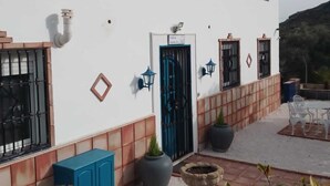 A white building with a blue door, a small garden with potted plants, and outdoor furniture. - Casa Camino (Valle de Abdalagis)