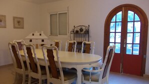 A dining area with a round table and six chairs, a window with blinds, a door with glass panels, and a wall-mounted shelf with books. - Casa Camino (Valle de Abdalagis)