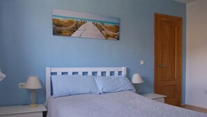 A bedroom with a bed, two bedside tables with lamps, a wooden door, and a wall-mounted picture of a beach pathway. - Casa Camino (Valle de Abdalagis)