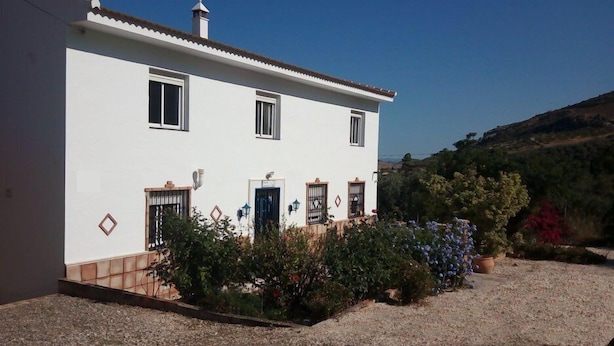 A two-story building with a white facade, multiple windows, and a small garden area with various plants. - Casa Camino (Valle de Abdalagis)