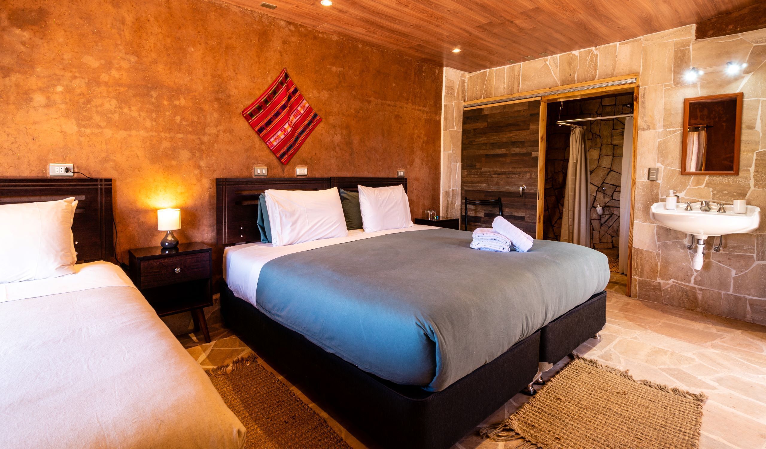 A hotel room with a large bed, a nightstand, a lamp, a bathroom with a sink and mirror, and a stone wall.