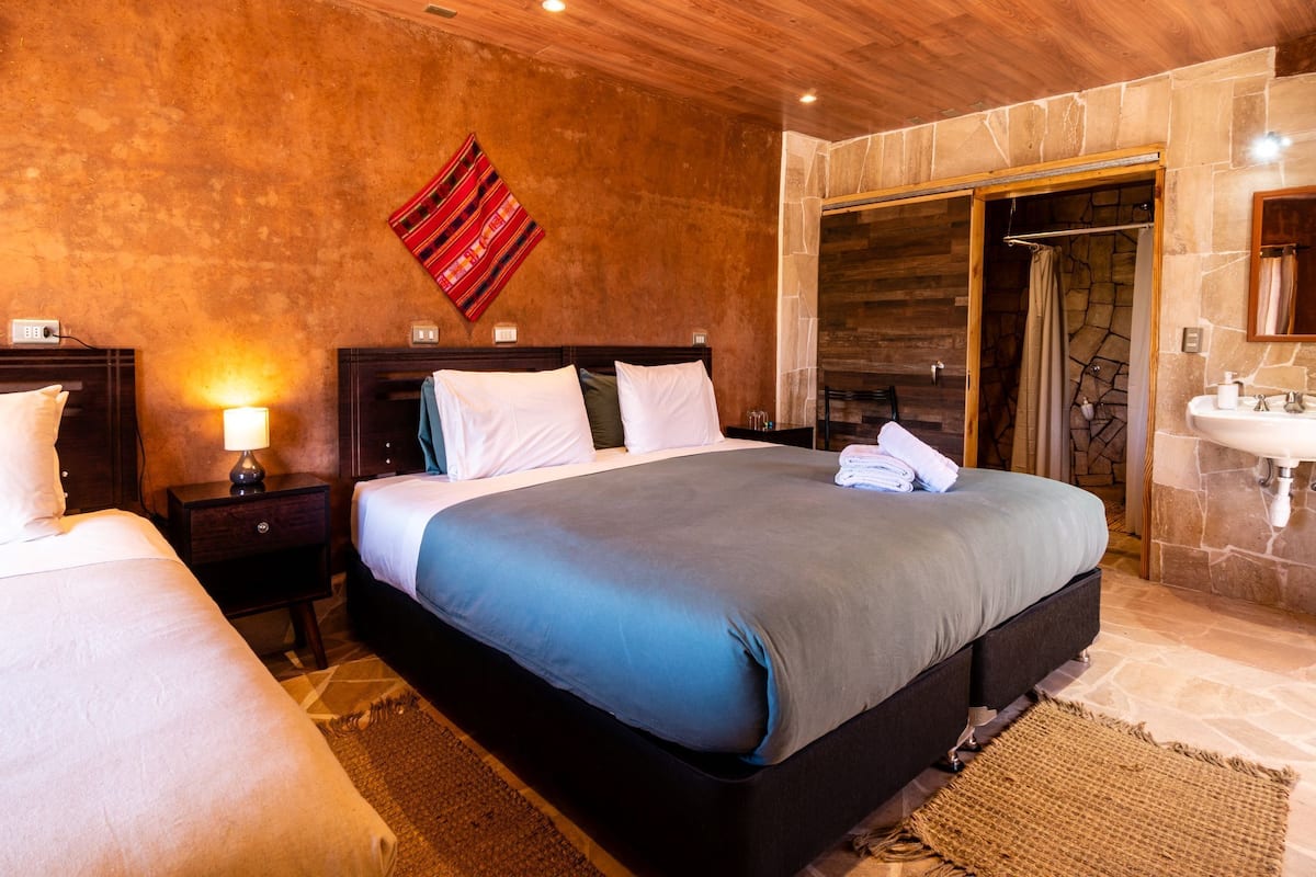 A hotel room with a large bed, a nightstand, a lamp, a bathroom with a sink and mirror, and a stone wall.
