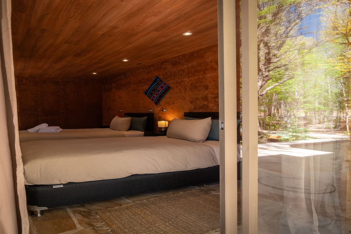Une chambre avec deux lits, un plafond en bois, un tableau accroché au mur et une vue sur une forêt, visible à travers une porte vitrée.
