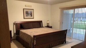Un dormitorio con cama de madera, mesita de noche, lámpara, un cuadro en la pared y vistas por la ventana.