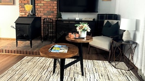 Ein gemütliches Wohnzimmer mit Kamin, Flachbildfernseher, einem kleinen runden Tisch mit Zeitschriften, einer Couch und einer Lampe.