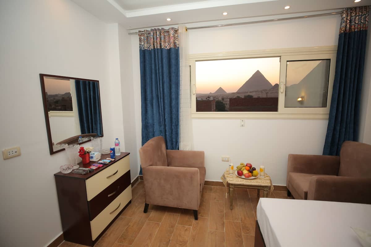 A hotel room with a bed, two armchairs, a small table with fruit and a bottle, a dresser, a mirror, and a window with a view of a pyramid.