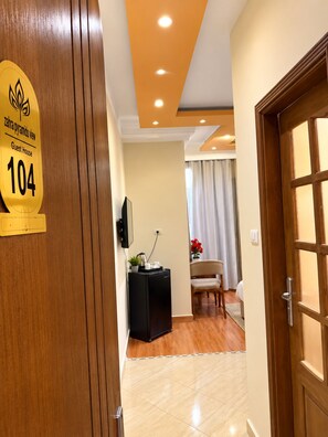 Habitació d'hotel amb una porta de fusta, un senyal que indica que és una casa d'hostes, una petita taula negra amb una paperera, una cadira i un llit amb un llençol blanc.