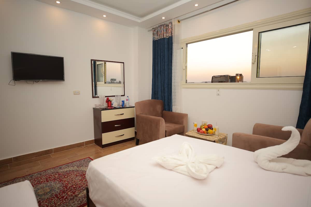 A hotel room with a bed, two chairs, a TV, a mirror, a window with curtains, and a small table with fruit and candles.