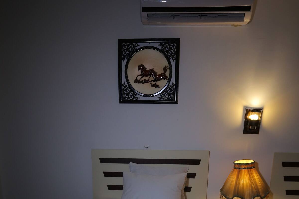 A hotel room with a bed, a wall-mounted air conditioner, a framed picture, a bedside lamp, and a decorative towel arrangement.