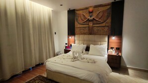 Habitación de hotel con una cama grande, cabecera con diseño de temática egipcia, mesita de noche con lámpara y un gran ventanal con cortinas.