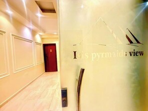Un pasillo con una puerta roja al final, un muro de cristal con el texto 'It's pyramids view' y una lámpara moderna en el techo.