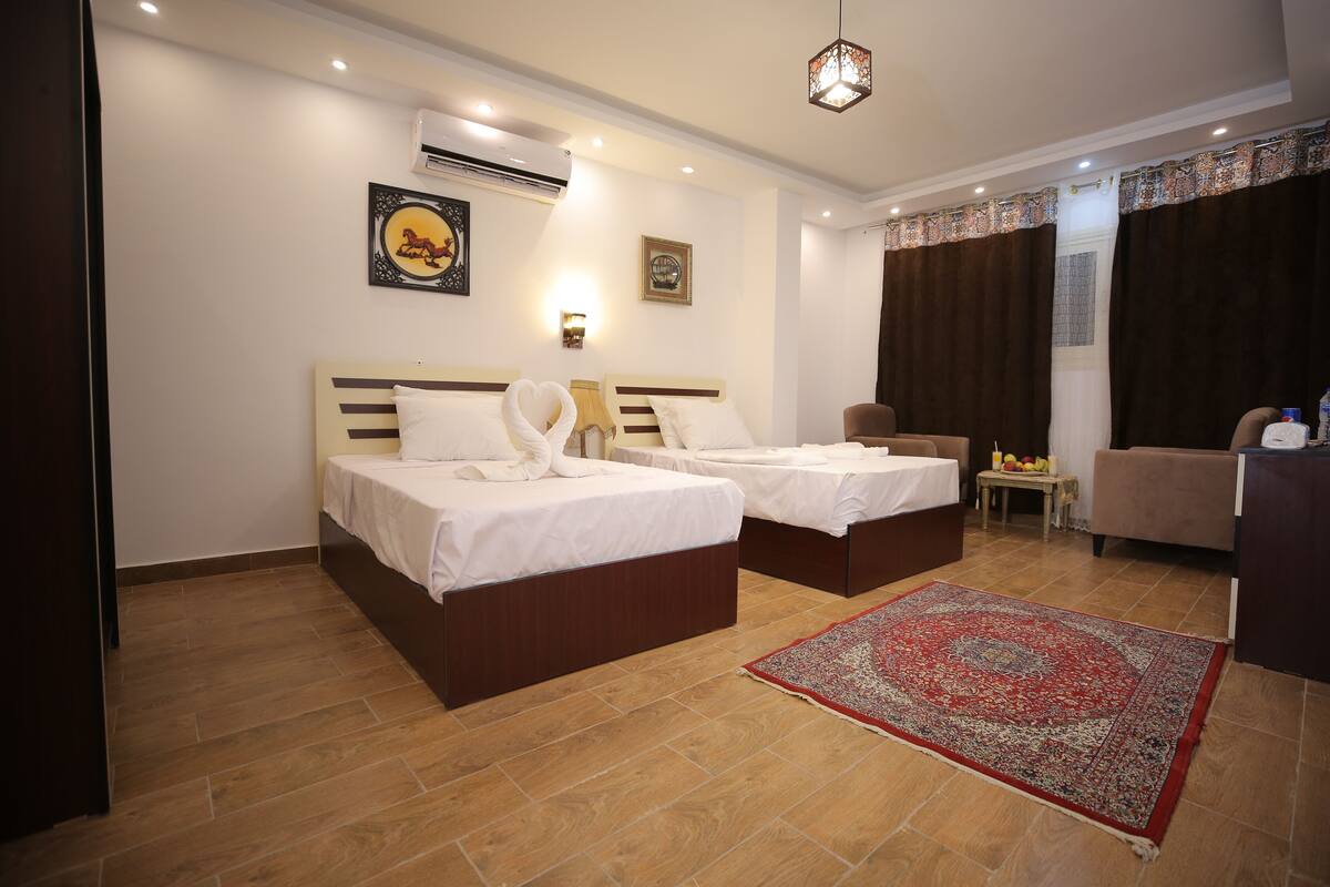 A hotel room with two beds, a red and white patterned rug, a small table with fruit, and a chair.