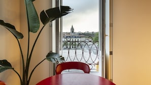 A room with a red table and a chair, a large plant, and a view of a building through a window with a metal railing.