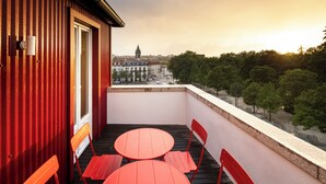 A balcony with red chairs and a table, overlooking a cityscape with buildings and trees.