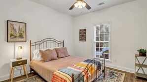 Une chambre à coucher comprenant un lit, une table de chevet, un ventilateur de plafond, une fenêtre avec des stores, un tableau encadré au mur et une petite plante posée sur une table d’appoint.