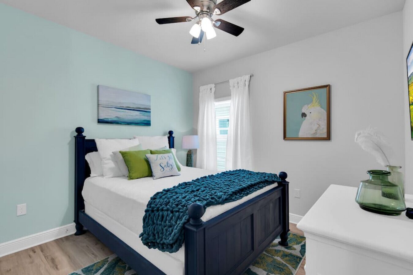A bedroom with a bed, a ceiling fan, a painting, a window with curtains, and a bedside table with a vase.
