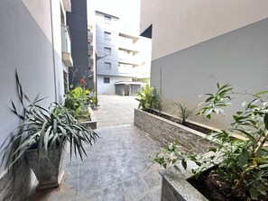 A narrow corridor with tiled flooring, potted plants, and a view of a multi-story building in the background.