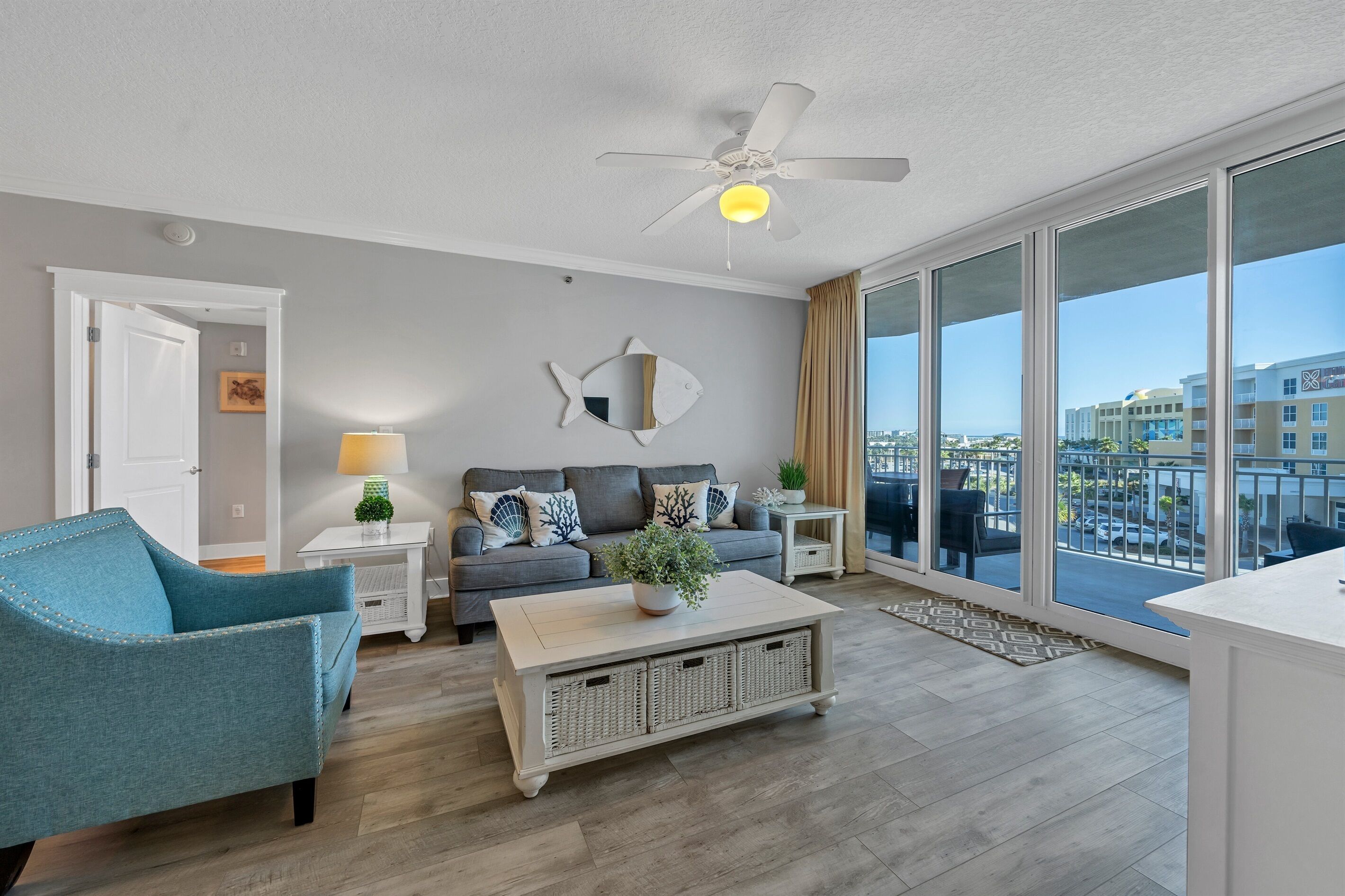 A modern living room with a sofa, a coffee table, a blue armchair, a ceiling fan, and a large window with a city view.
