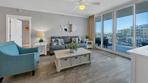 A modern living room with a sofa, a coffee table, a blue armchair, a ceiling fan, and a large window with a city view. - EscapetoWaterscape-Family Friendly 2/2.5 condo (Fort Walton Beach)