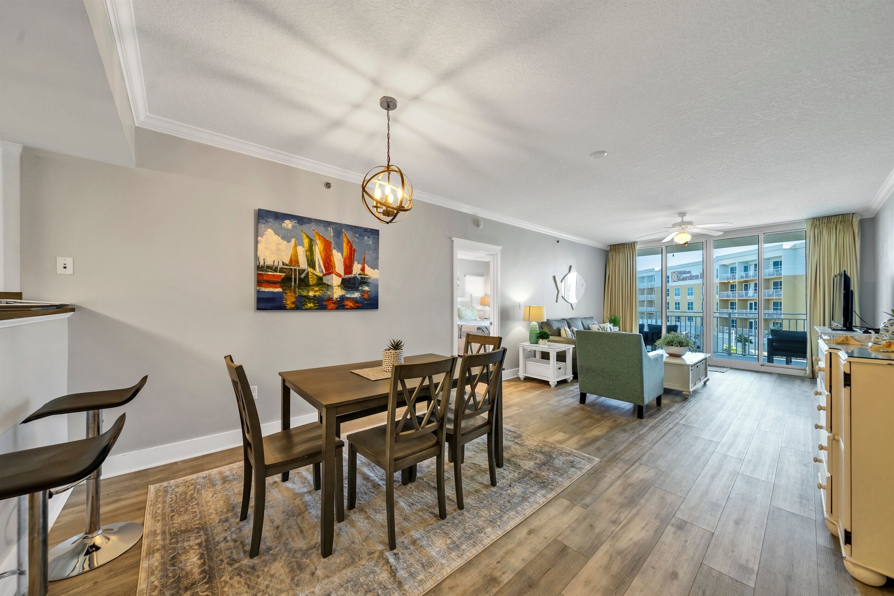 A modern living room with a dining area, a painting on the wall, and a view through large windows.
