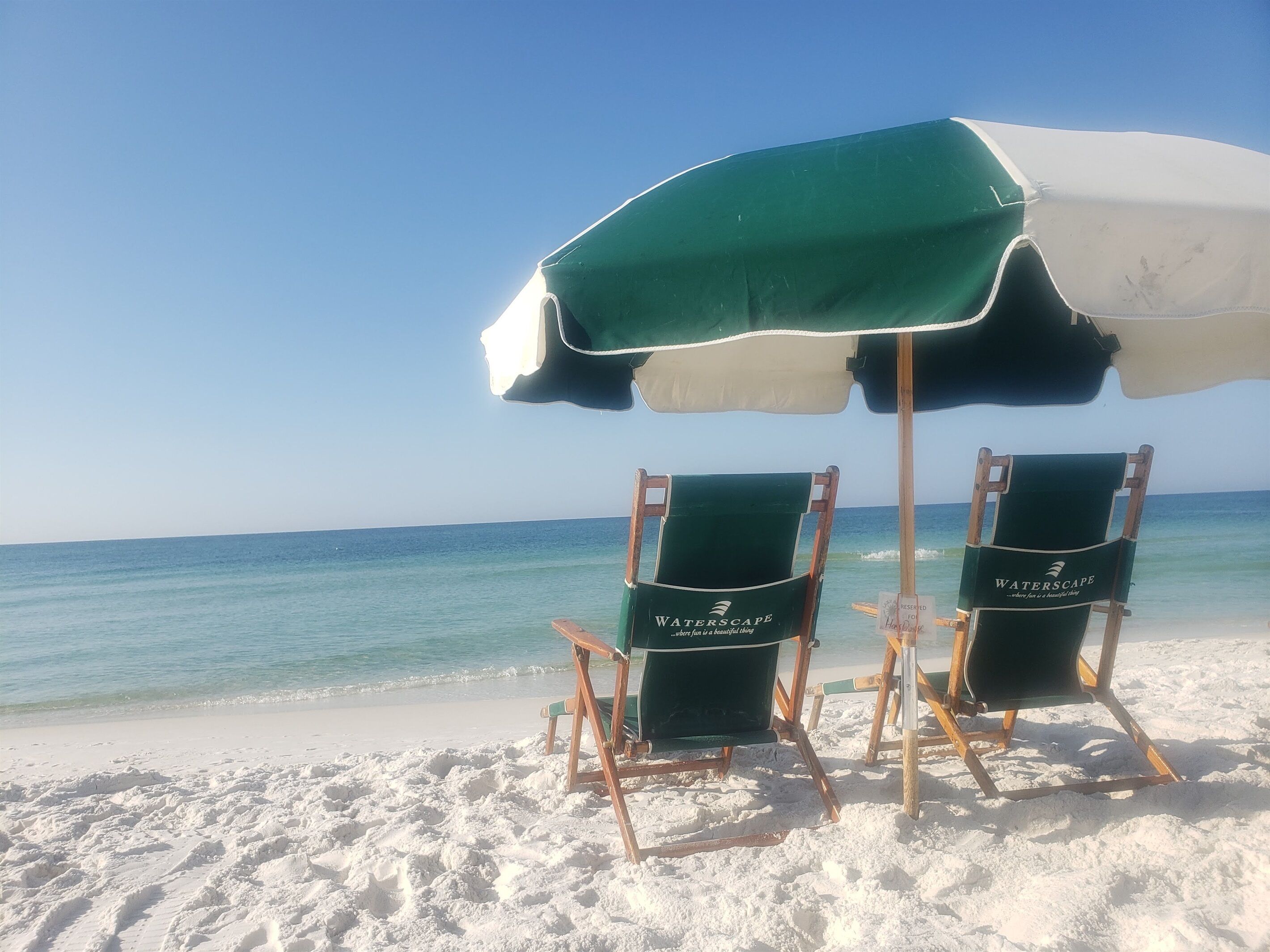 Two beach chairs under a green and white umbrella on a sandy beach with a clear view of the ocean.